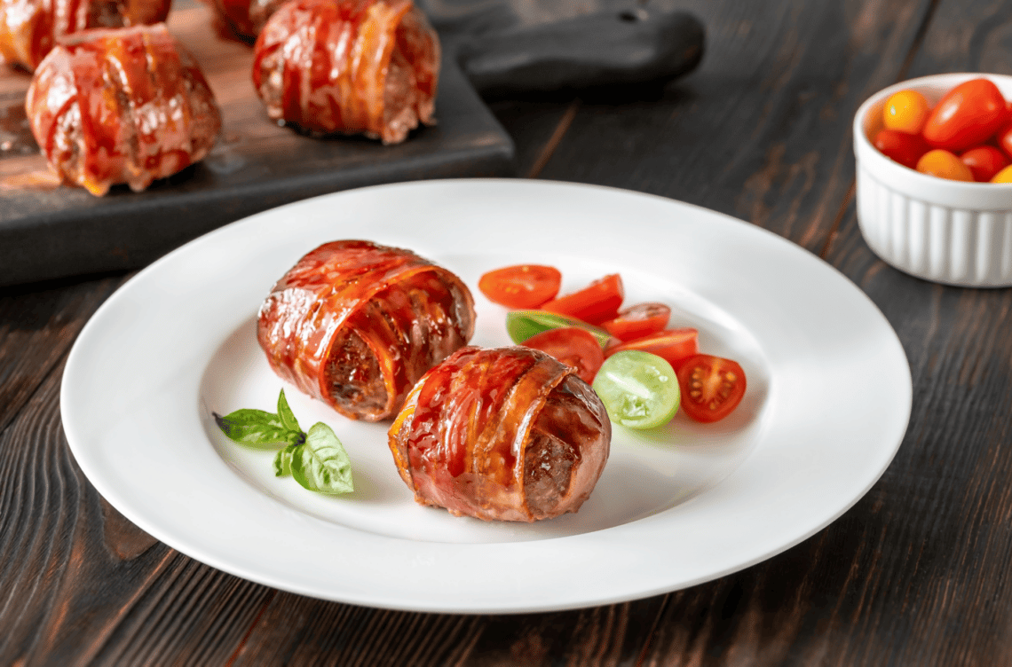 2 bacon wrapped paleo meatballs on a white plate with sliced cherry tomatoes