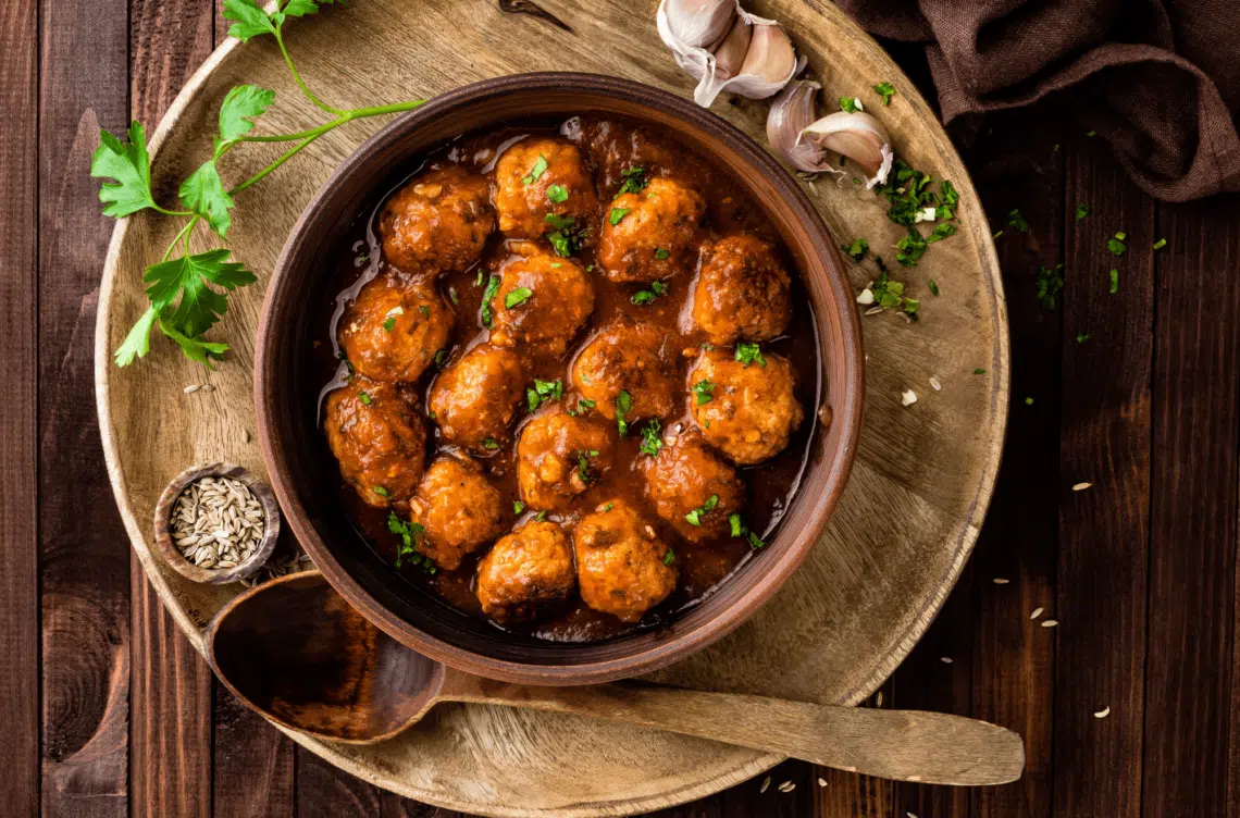 Saucy Asian Meatballs Appetizer In A Wooden Bowl With Garlic, Sesame Seeds, And Wooden Spoon On The Side