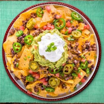 The Best Mexican Chorizo Nachos On A Round Serving Plate With Sour Cream On Top